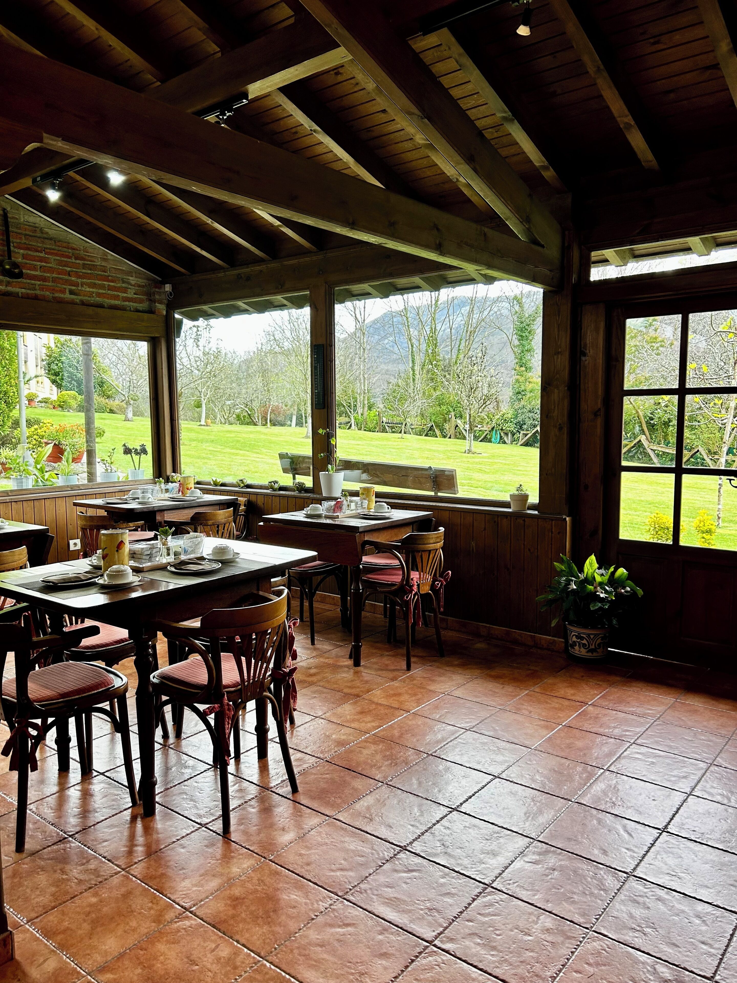 A dining area with wooden tables and chairs, a view of a garden, and a brick wall.