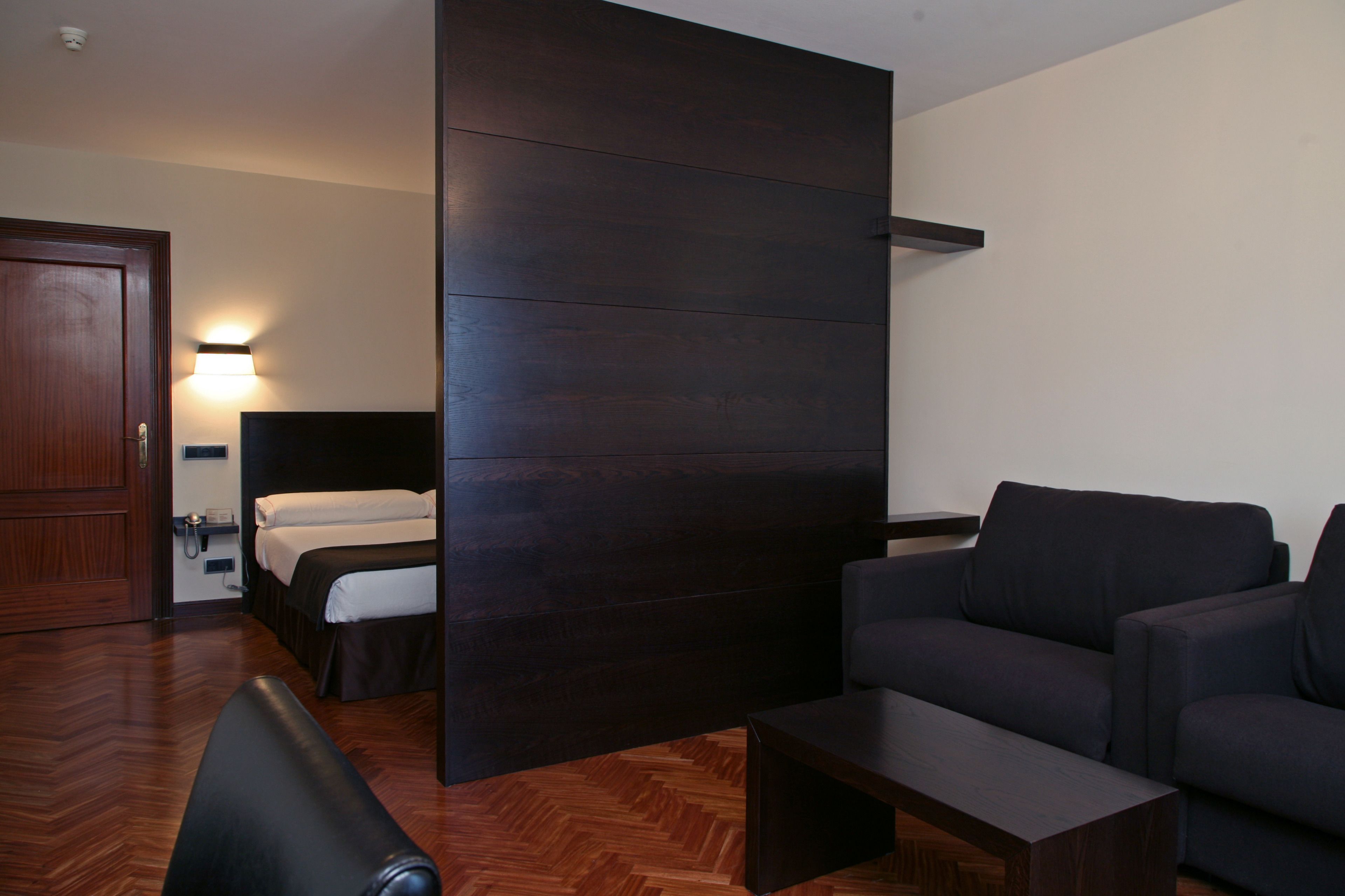 Una habitación de hotel moderna con suelo de madera, cama con ropa blanca, sofá oscuro, una mesita de centro pequeña y un televisor montado en la pared.