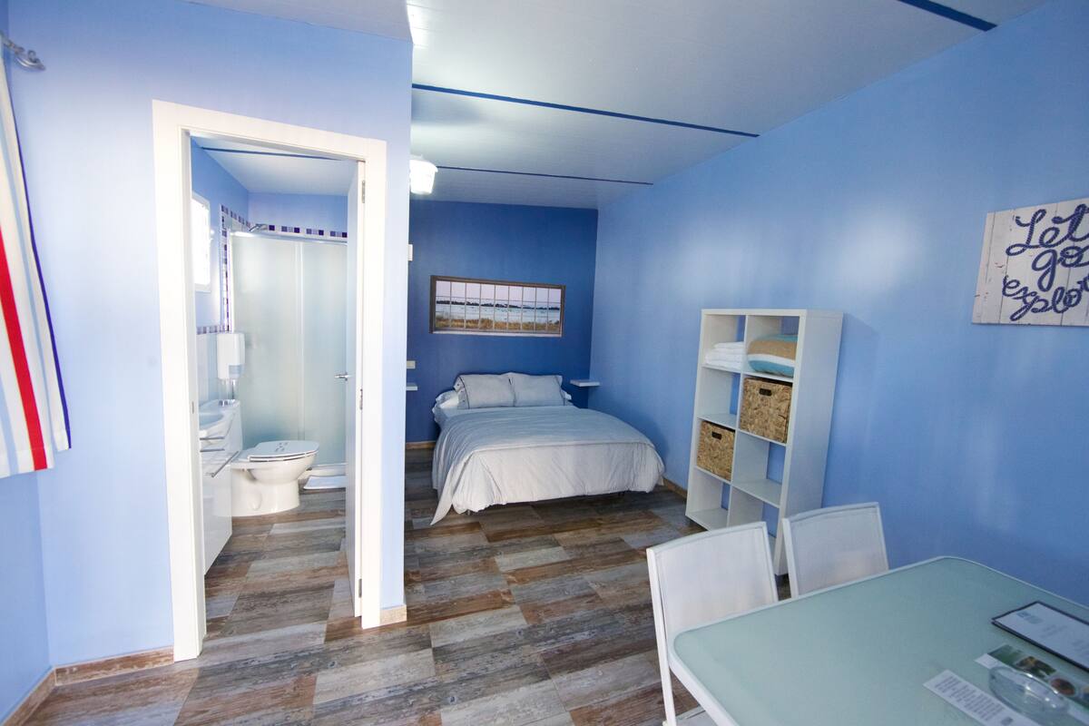 A bedroom with a bed, a desk, and a chair. There is a bathroom visible in the background.