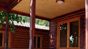 Una cabaña de madera con un porche cubierto, una mesa con sillas y un ventanal con vistas a los árboles circundantes.
