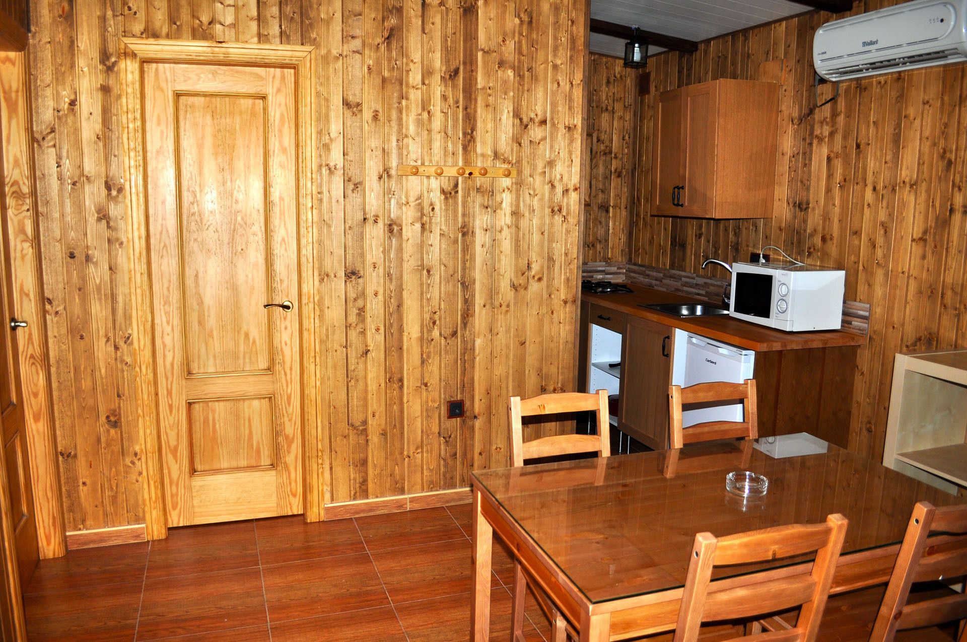 A wooden-paneled room with a wooden door, a kitchen area including a microwave and a dishwasher, and a dining table with chairs.