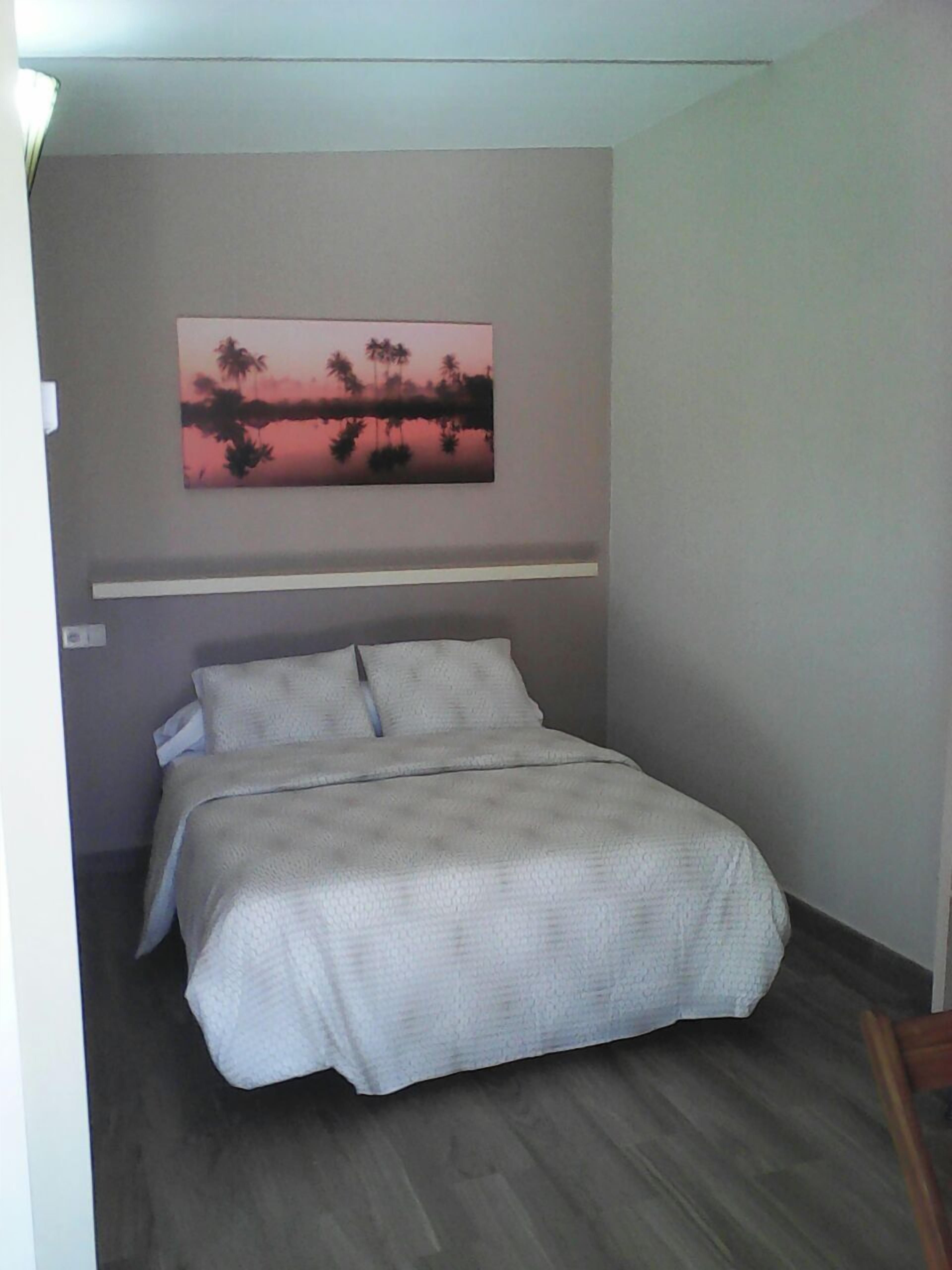 A bedroom with a bed, a painting of a sunset, and a wooden floor.