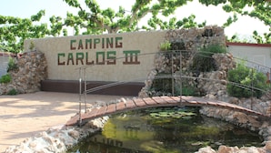Un muro de piedra con señalización 'CAMPING CARLOS III', un pequeño puente sobre un estanque y vegetación circundante.