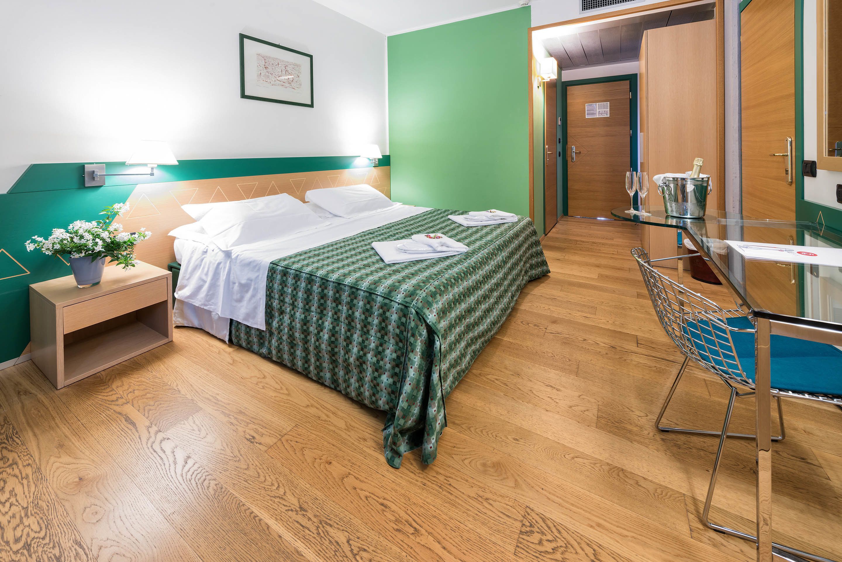 A hotel room with a bed, a bedside table with a lamp, a small potted plant, a glass table with a chair, and a green wall.