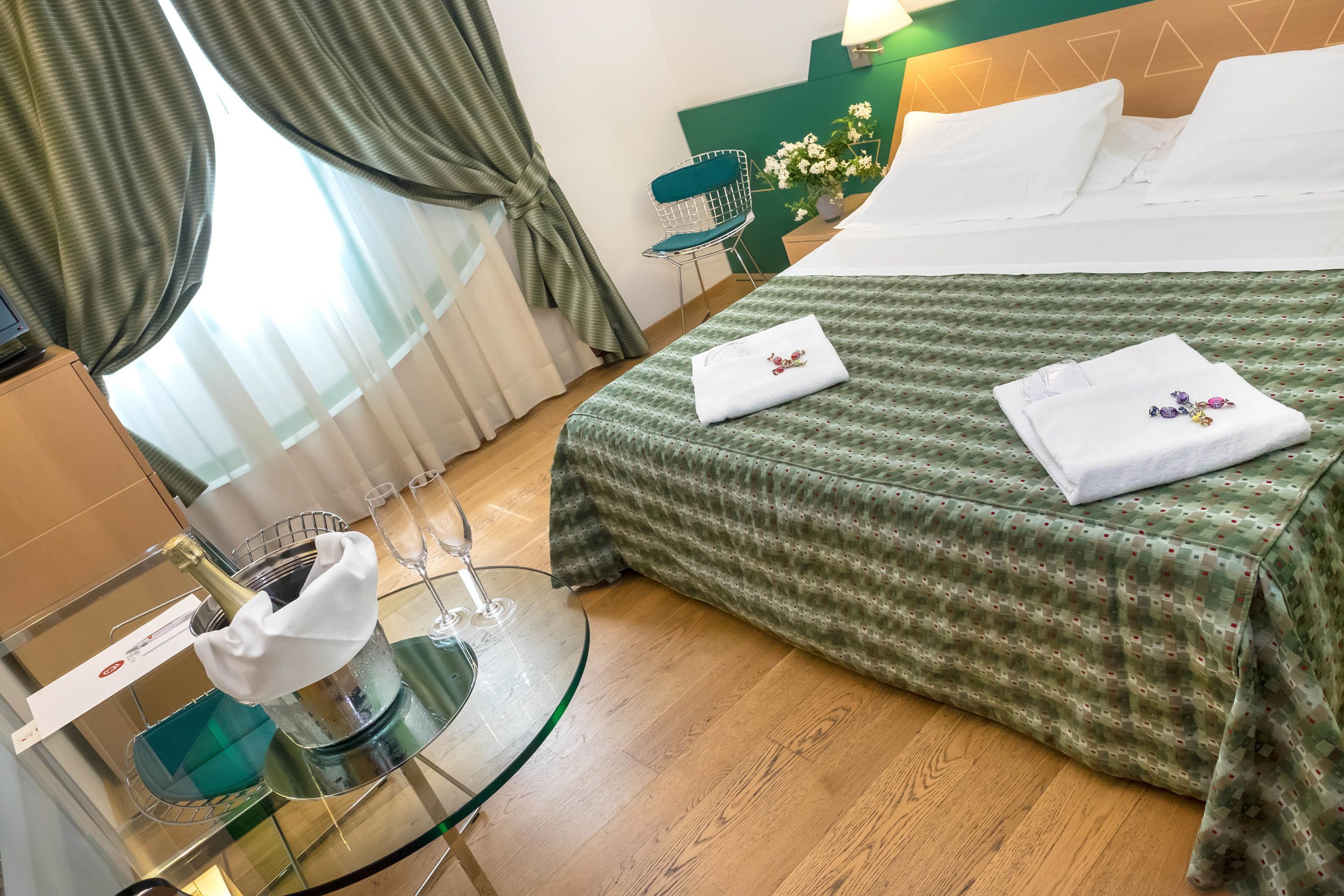 A hotel room with a bed, a glass table with a bottle, a chair, and a green wall.