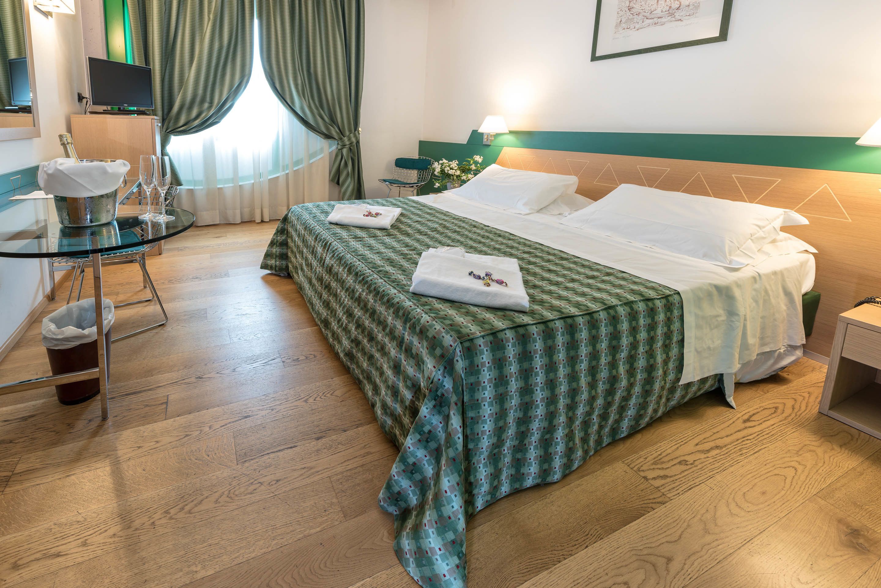 Quarto de hotel com cama, mesa de cabeceira, televisão e uma pequena mesa com garrafa e copos.