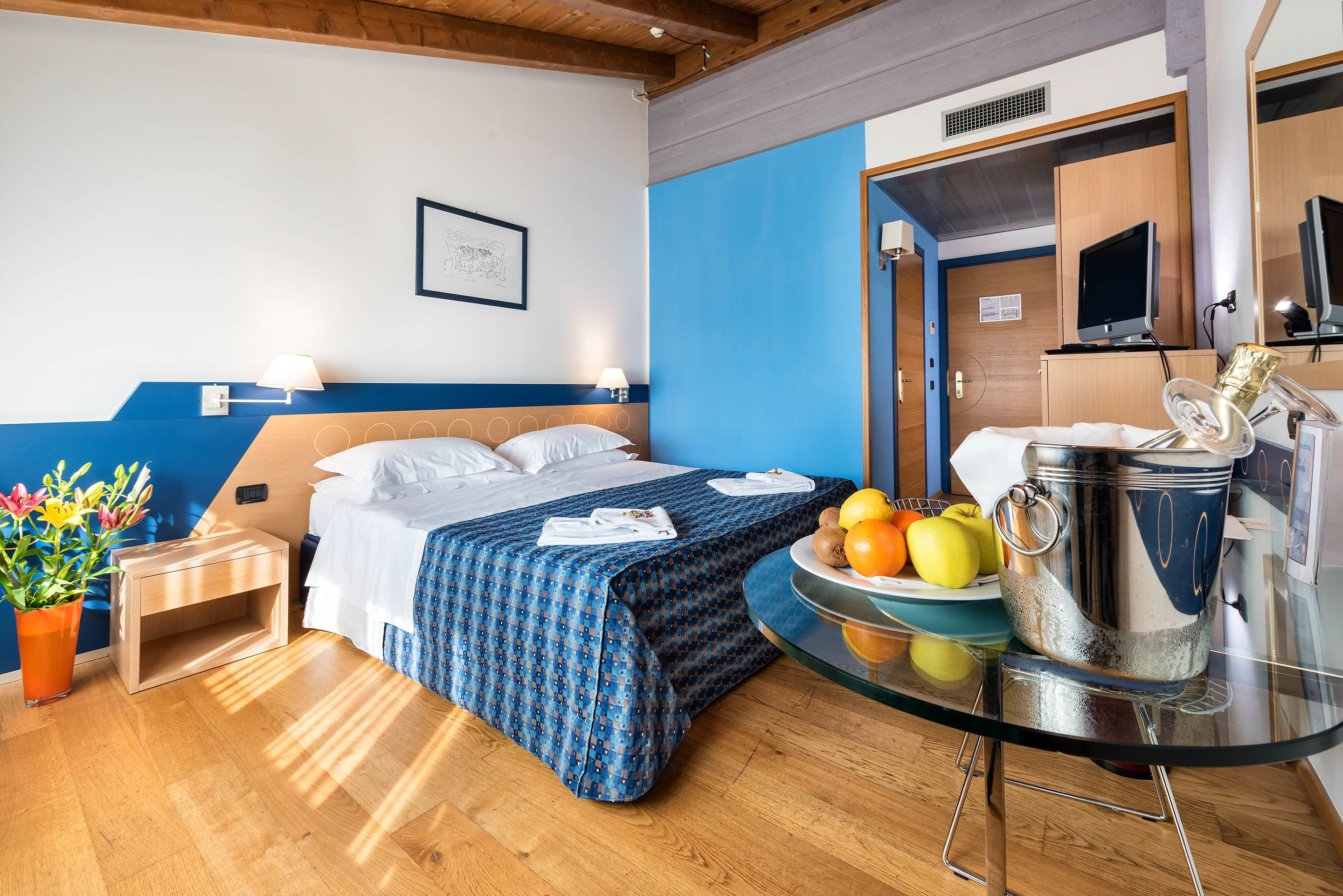 A hotel room with a bed, bedside table, lamp, fruit bowl, and a kitchen area with a stove.