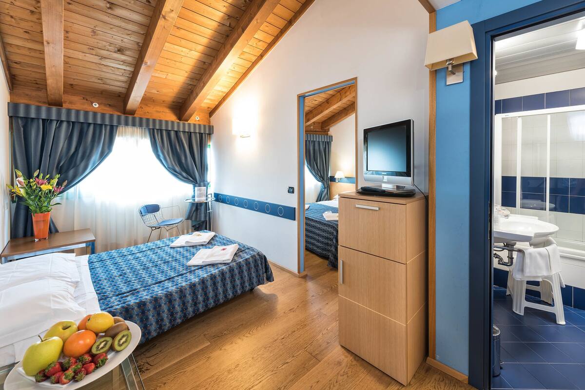 Habitación de hotel con dos camas, un escritorio, un televisor, un baño pequeño y una bandeja de desayuno con frutas.