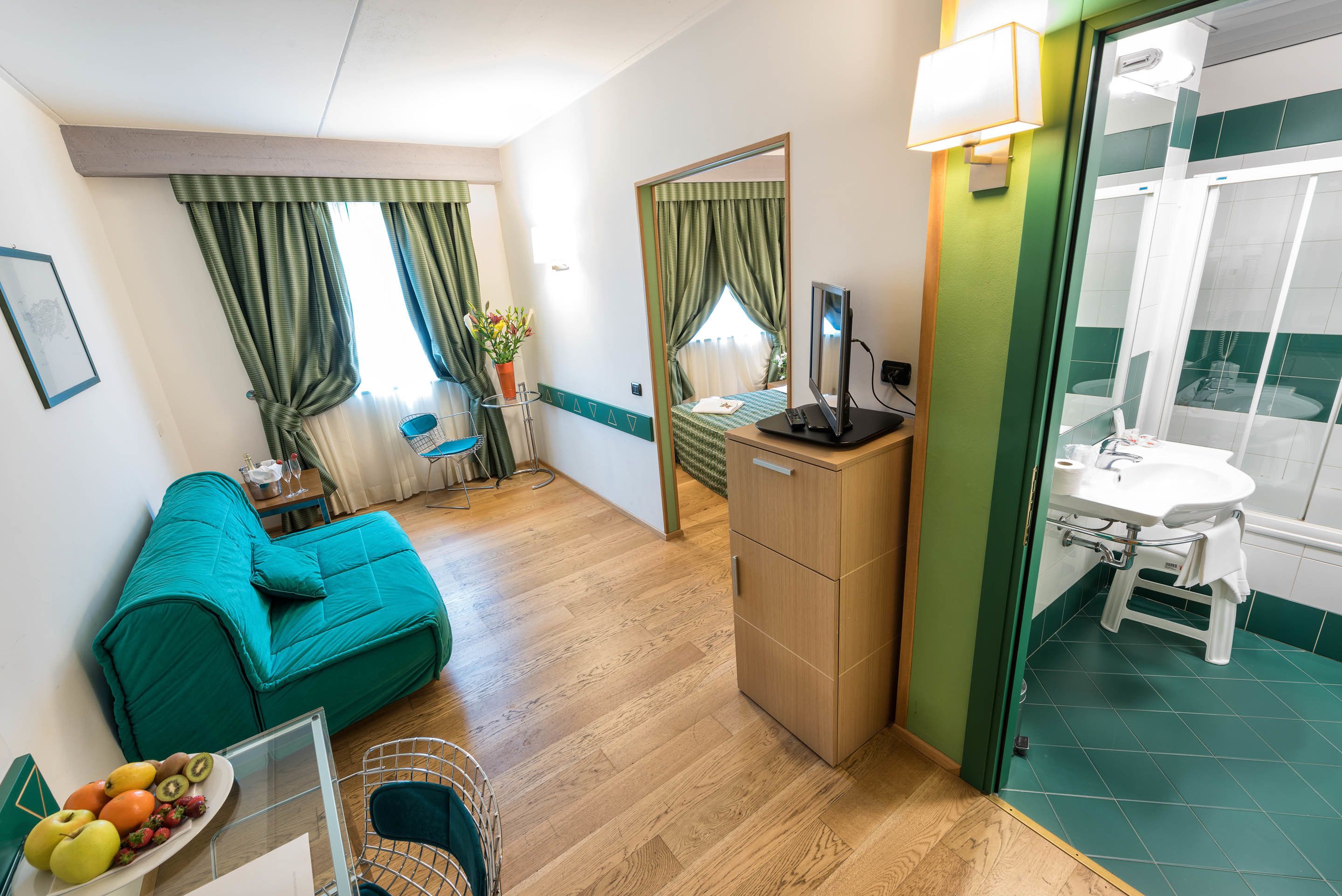 A compact hotel room with a teal sofa, a wooden desk, a small dining table with fruit, a mirror, and a bathroom with a green-tiled shower.