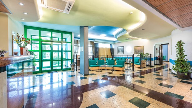 A spacious lobby with a checkered floor, green and blue seating areas, and large windows. - West Point Airport Hotel (Villafranca di Verona)