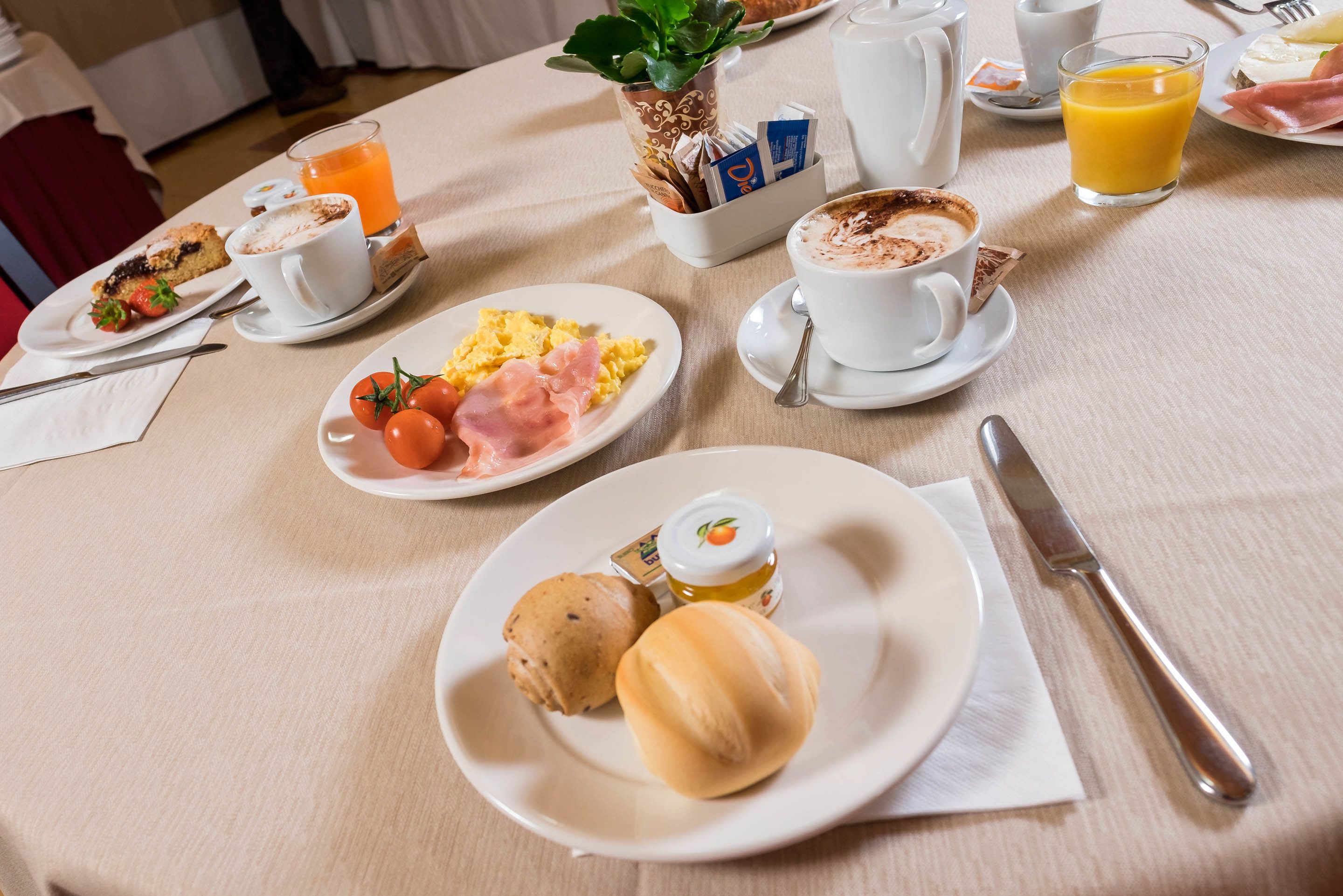 A breakfast table with plates of eggs, tomatoes, ham, and bread rolls, along with cups of coffee and orange juice.
