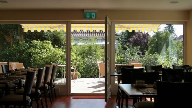An indoor dining area with tables and chairs, a glass door leading to an outdoor patio, and a yellow awning. - Parkhotel Lindau (Lindau)