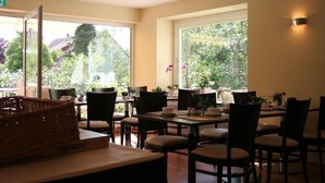 A dining area with multiple tables and chairs, large windows with a view of greenery, and a basket on the table. - Parkhotel Lindau (Lindau)