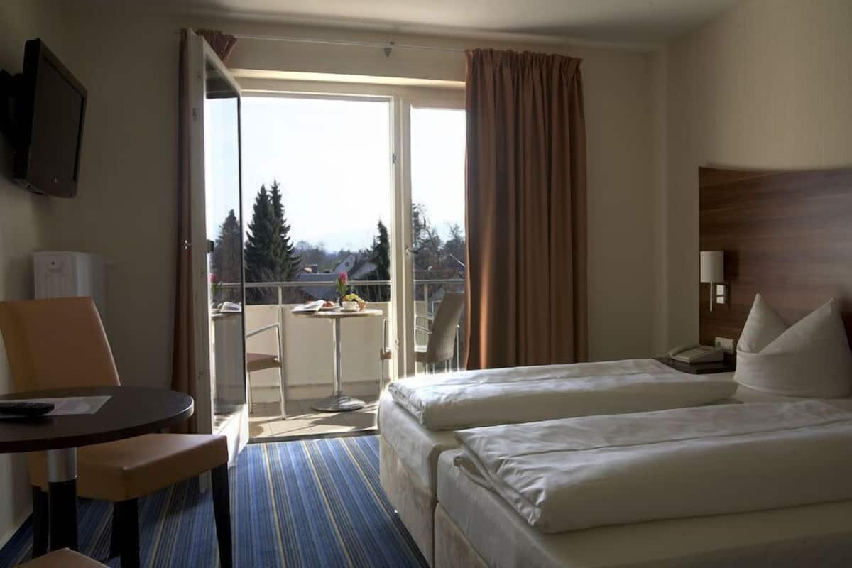 A hotel room with two beds, a small table, a chair, a TV, and a balcony with a view of trees and buildings.