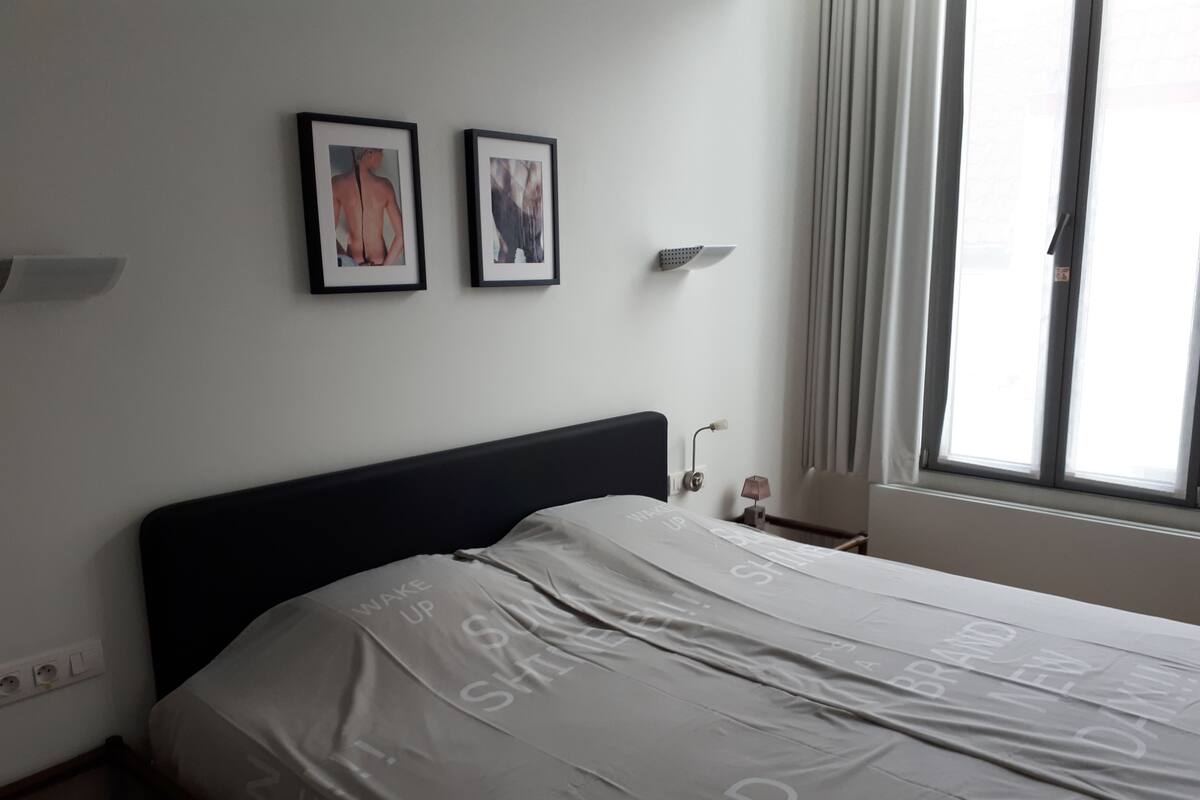 A bedroom with a bed, a nightstand, a wall-mounted lamp, and two framed pictures.
