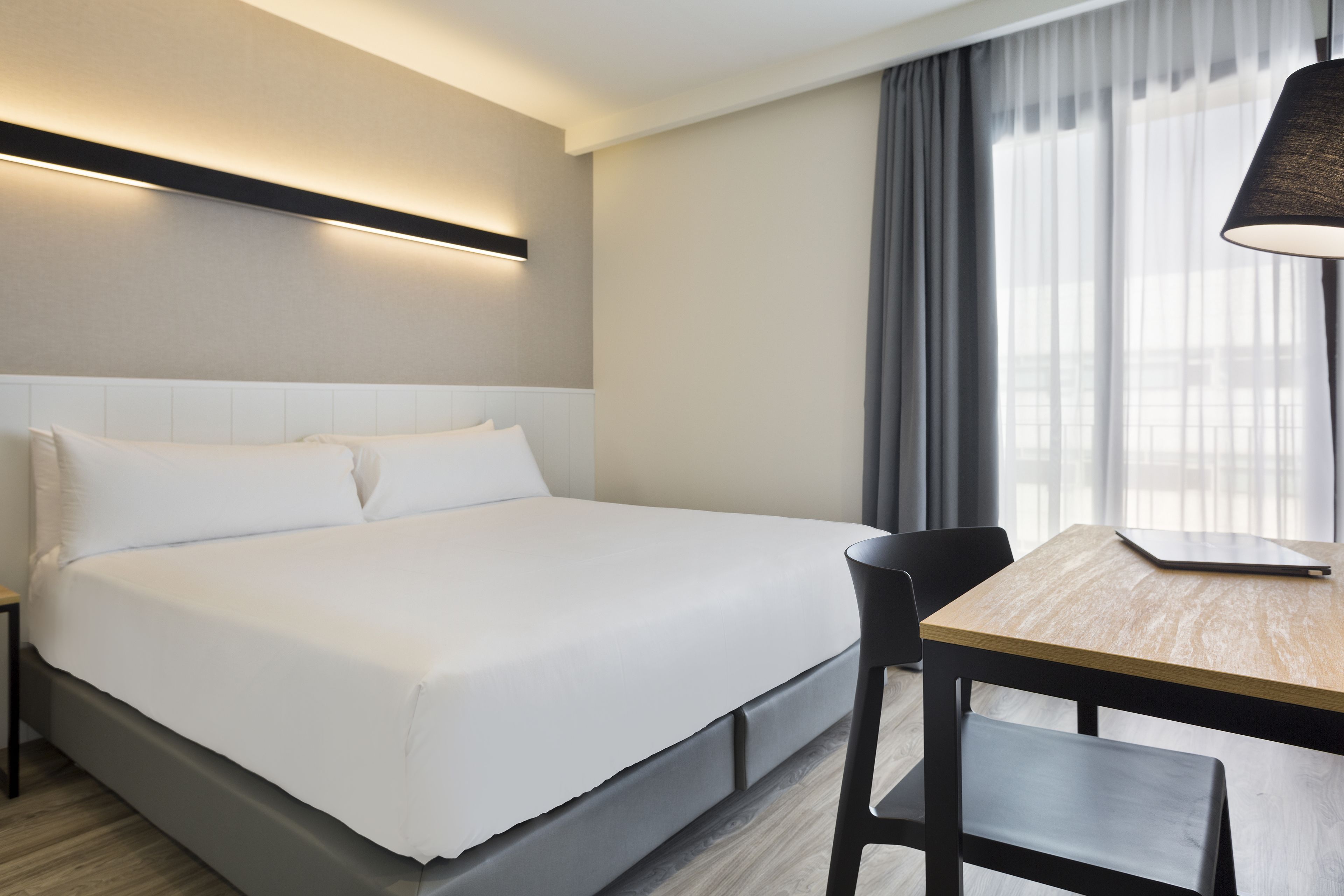 Quarto de hotel moderno com cama bem arrumada, escrivaninha de madeira com um laptop e uma cadeira.