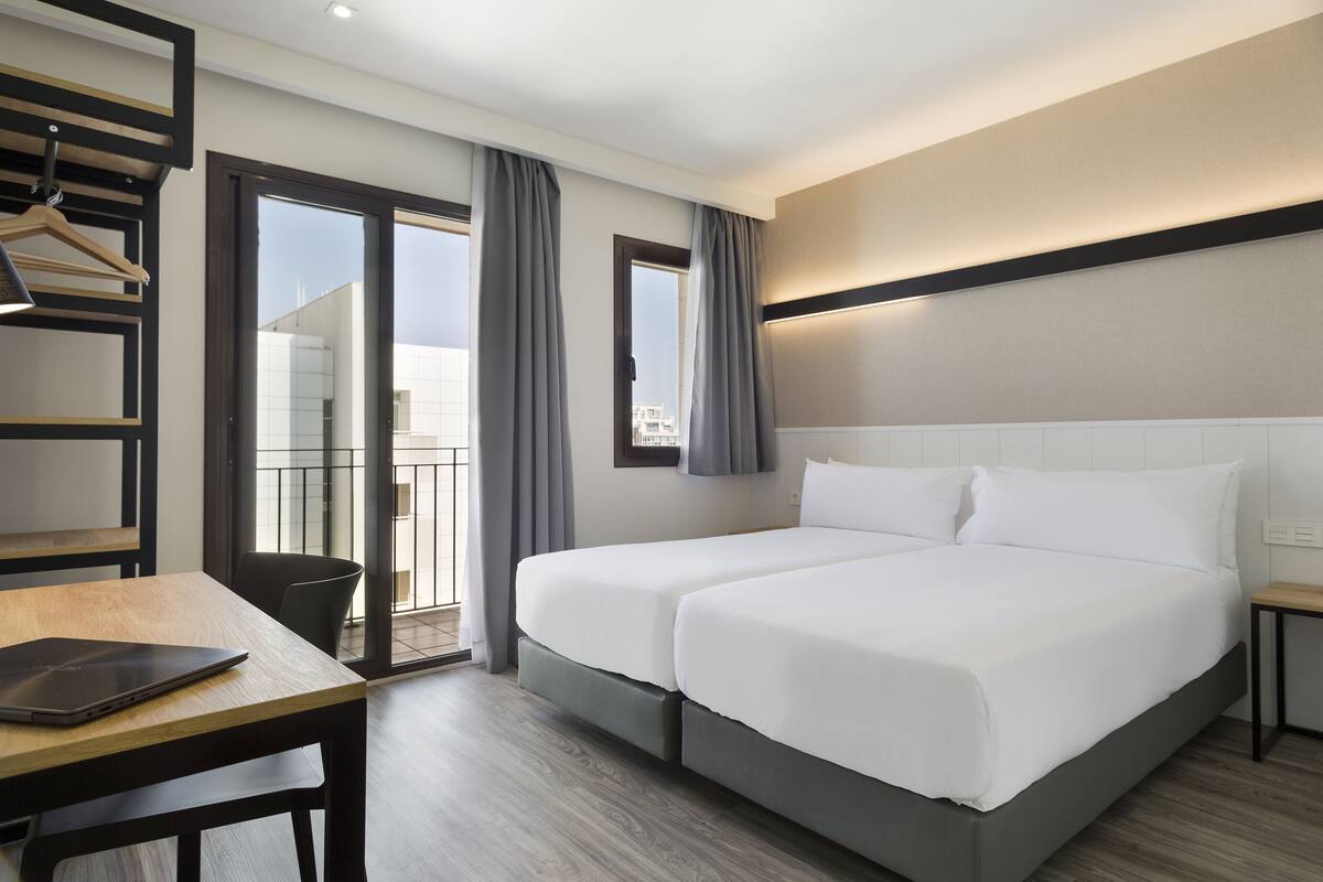 A hotel room with a double bed, a desk with a laptop, a chair, a small table, and a balcony with curtains.