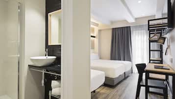 A modern hotel room with a glass shower, a white sink, a bed, a desk, and a TV mounted on the wall.