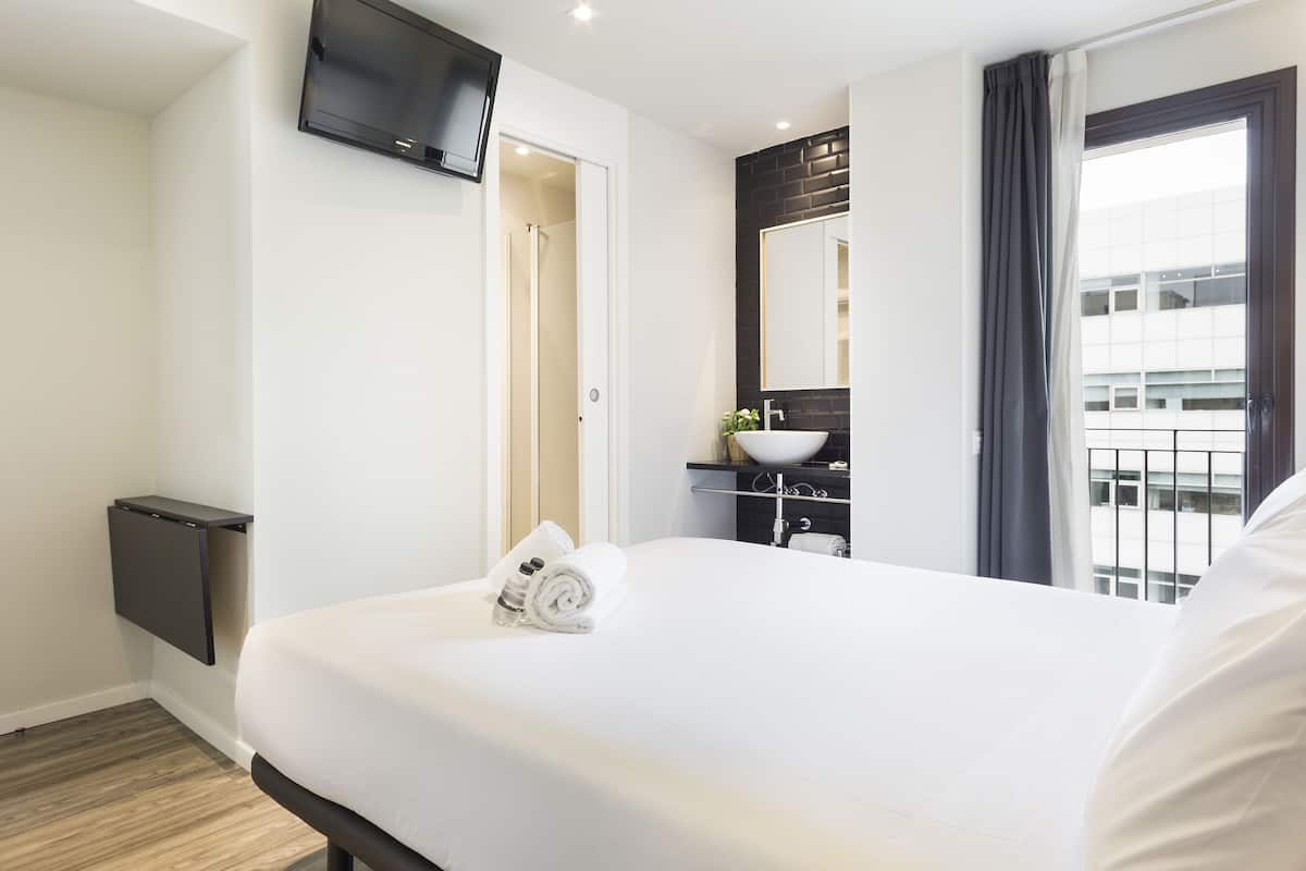 A hotel room with a large bed, a flat-screen TV mounted on the wall, a bathroom with a dark backsplash, and a window with curtains.