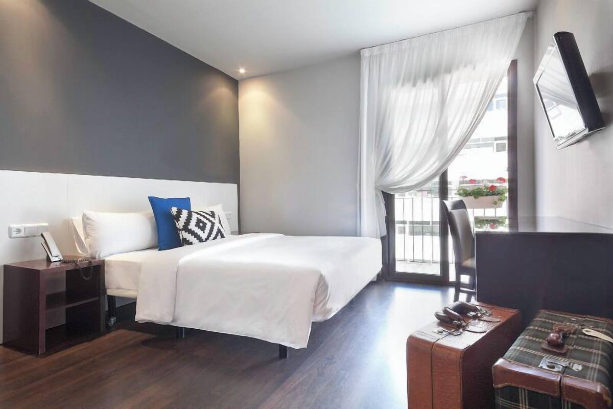 A modern hotel room with a bed, a desk, a chair, and a window with curtains.