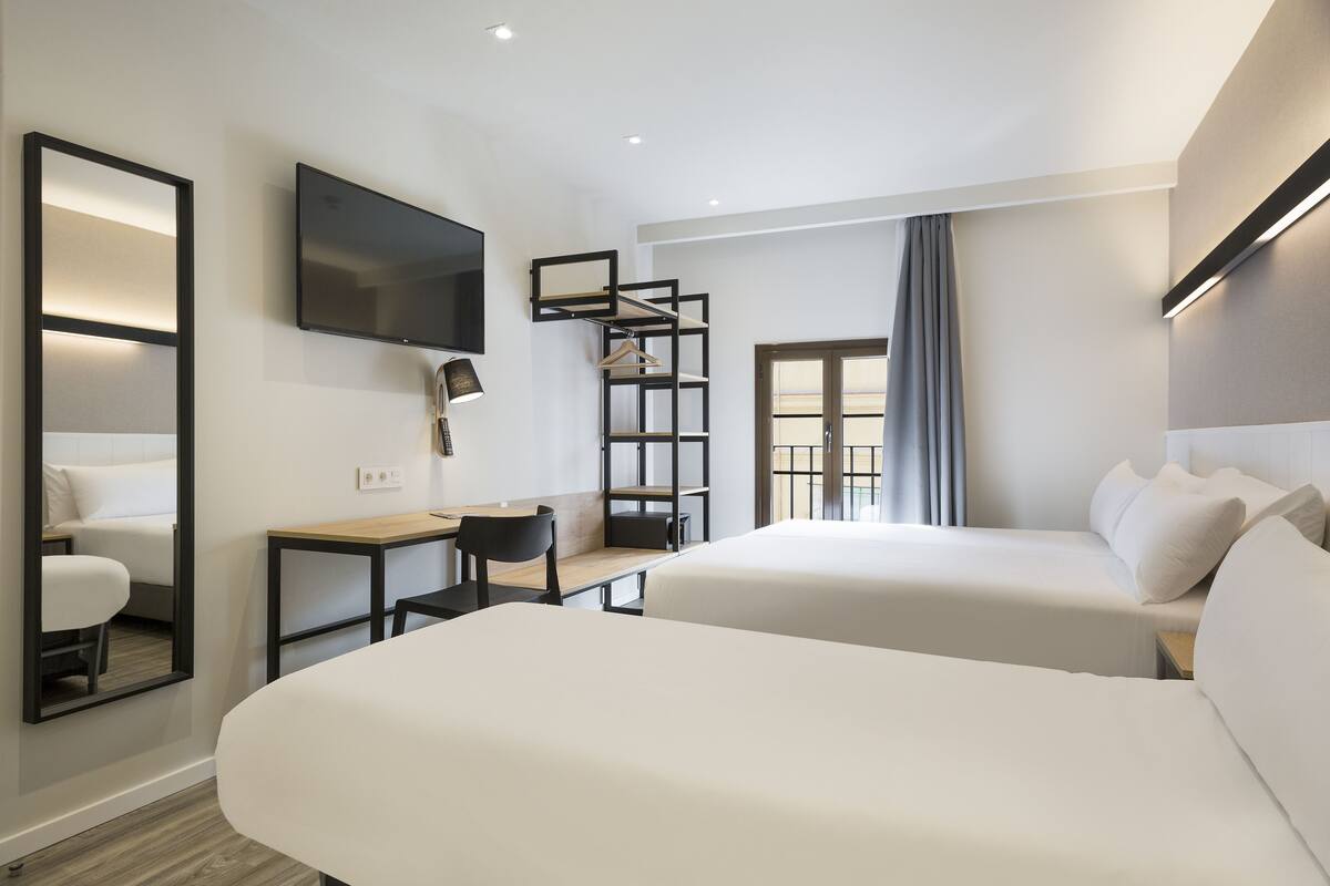 A modern hotel room with two beds, a desk, a chair, a TV, and a bookshelf.