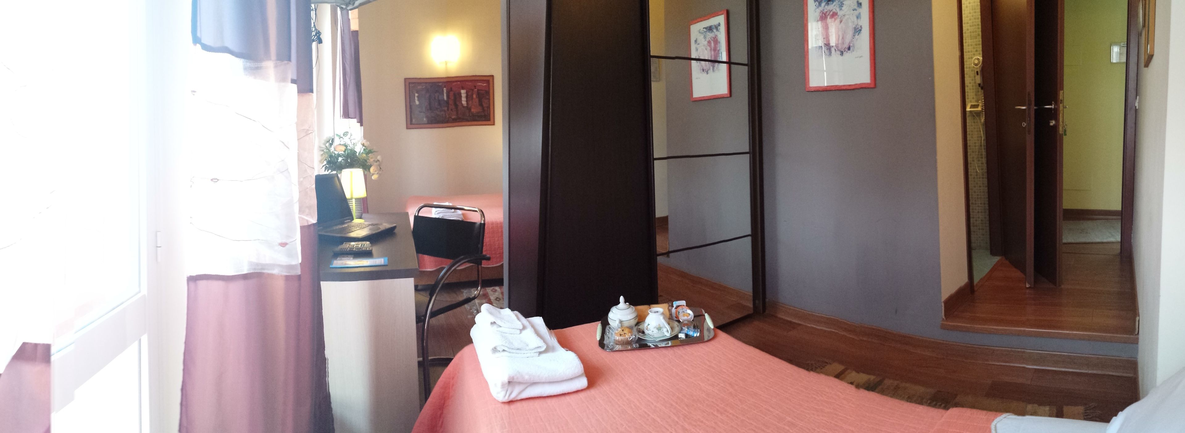 A hotel room with a bed, a table with a tray of toiletries, a chair, a desk with a laptop, and a window with curtains.