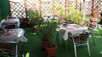 Area pranzo all'aperto con tavoli apparecchiati per due, circondata da piante in vaso e un recinto in legno a griglia.