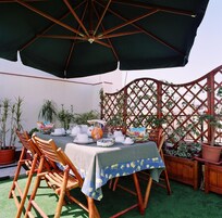 Uma área de refeições ao ar livre com uma mesa posta para quatro pessoas, rodeada por cadeiras de madeira e um guarda-sol preto.