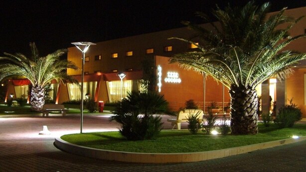 A well-lit hotel entrance with palm trees, a paved walkway, and a building with illuminated signage. - Hotel Cecere (Mottola)