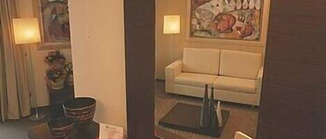 Habitación de hotel con sofá, un cuadro, lámpara y una mesa con una carpeta roja y una placa de identificación.