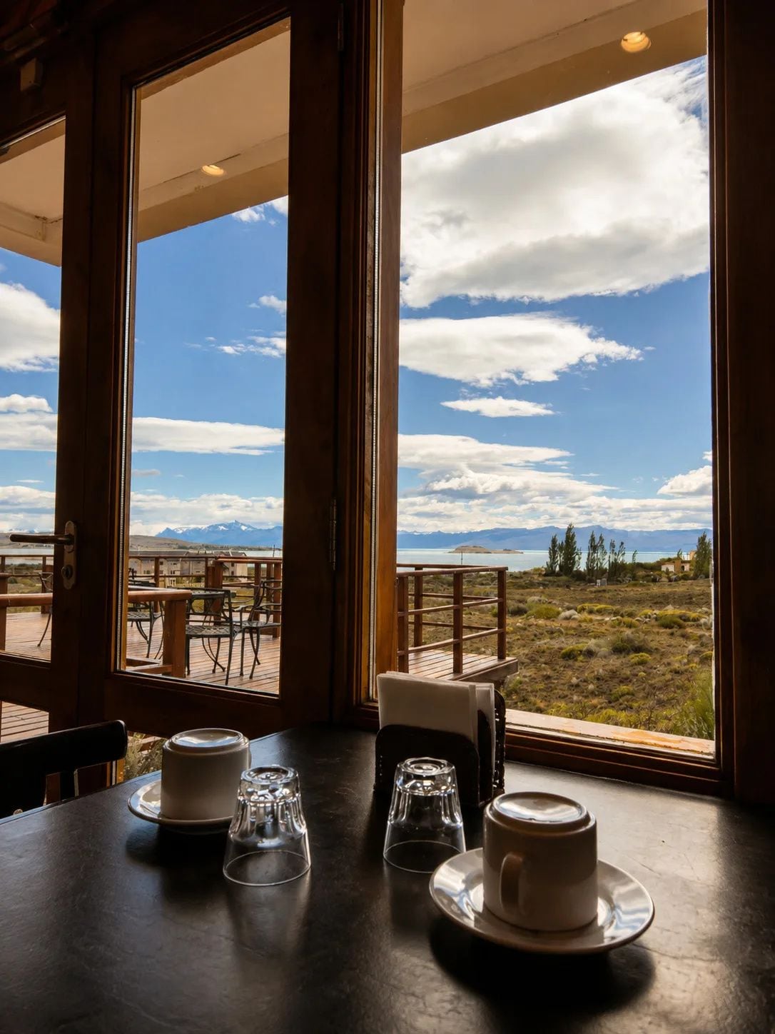 Una mesa de comedor con tazas y vasos, con vistas a un paisaje pintoresco que incluye montañas y una masa de agua.