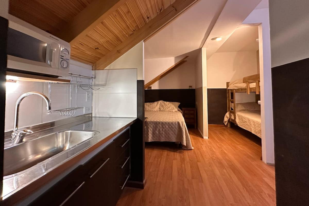 Cocina compacta con electrodomésticos modernos y techo de madera, que da paso a un dormitorio con literas.