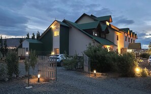 A multi-story building with a green roof and multiple windows, surrounded by a gravel driveway and a wooden fence. - Hostería Meulen (El Calafate)