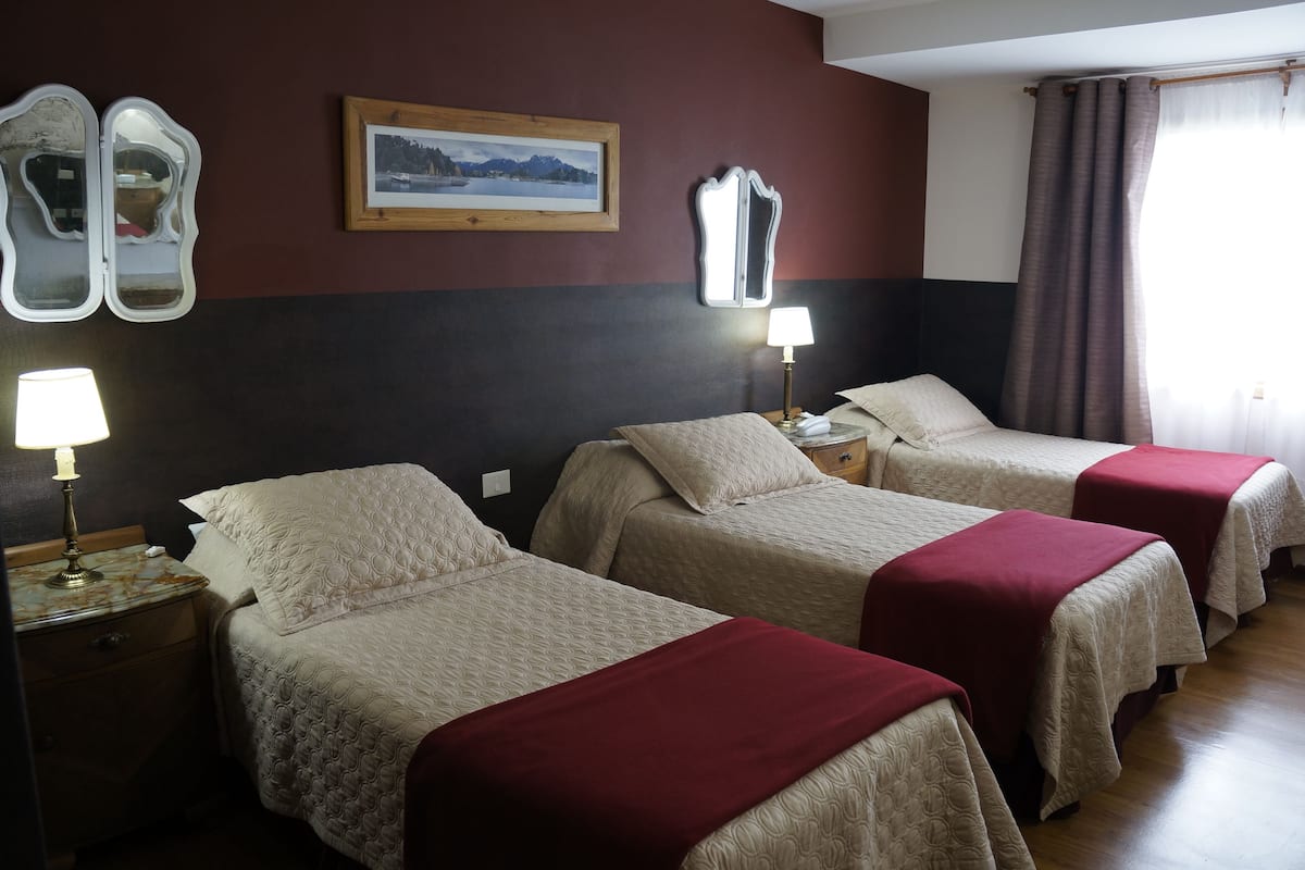 Habitación de hotel con tres camas, cada una con una manta roja, una lámpara de noche y un cuadro enmarcado en la pared.