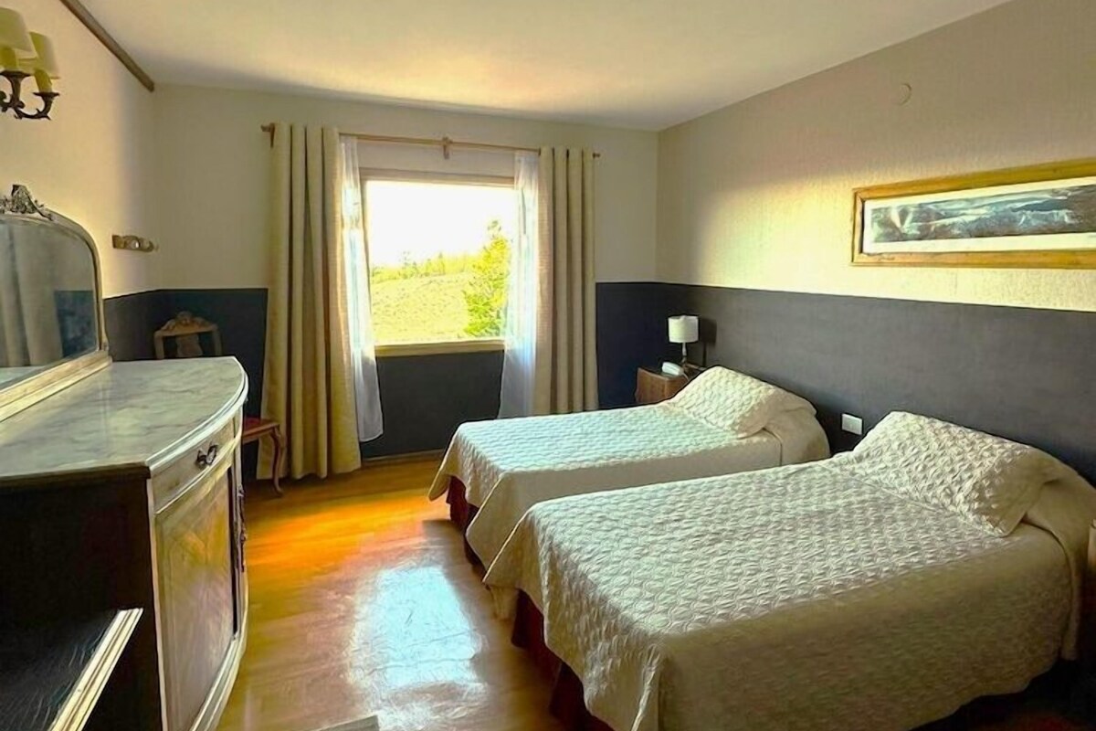 Une chambre d’hôtel avec deux lits, un bureau, une chaise, une fenêtre avec des rideaux et un tableau encadré au mur.
