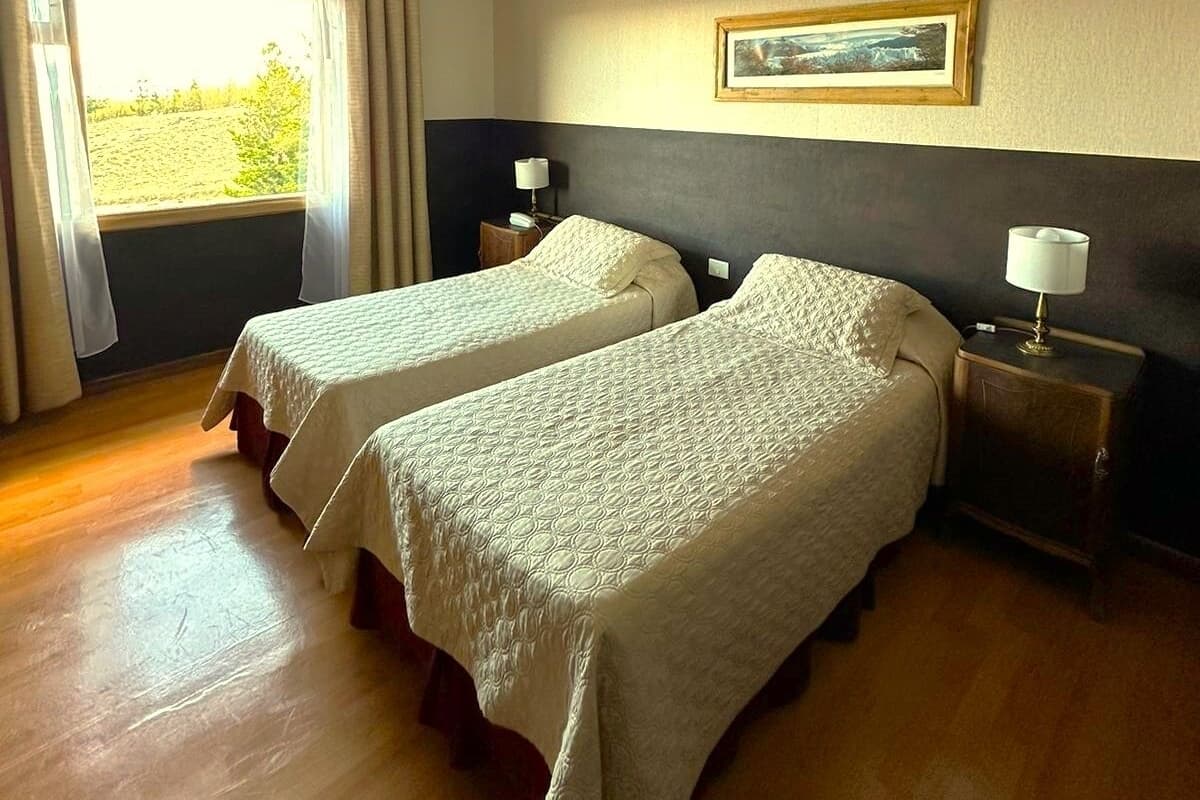 Una habitación de hotel con dos camas, una ventana con cortinas, una mesita de noche con lámpara y un cuadro en la pared.