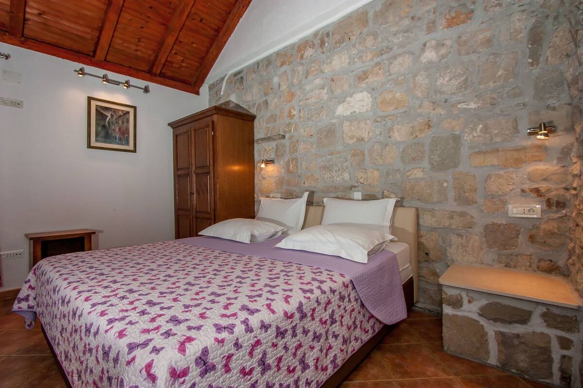A bedroom with a stone wall, wooden ceiling beams, a bed with a floral bedspread, a wooden wardrobe, and a bedside table.
