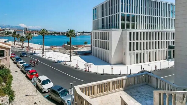 Un bâtiment moderne à la façade vitrée, un espace de stationnement avec des voitures et une vue dégagée sur une étendue d’eau.