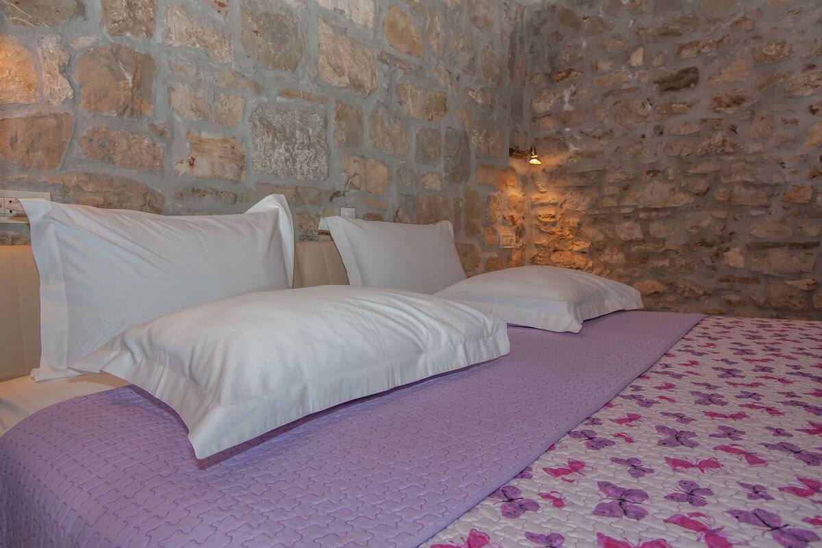 A bed with white linens and a purple blanket, set against a stone wall with a wall-mounted light.