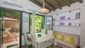 Un coin repas comprenant une table, des chaises et un canapé. On y trouve une fenêtre avec des rideaux, un mur en pierre et une étagère ornée d’objets décoratifs.