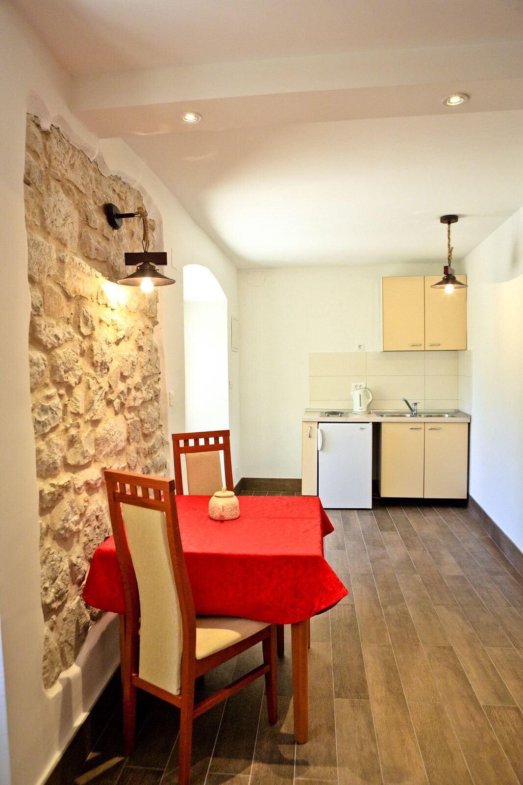 A dining area with a red table and chairs, a stone wall, a kitchenette with cabinets, and a ceiling light.