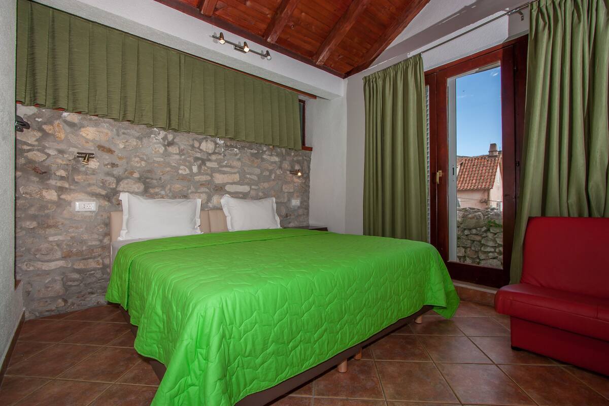 A bedroom with a stone wall, a bed with green bedding, a red chair, and a view through a glass door.