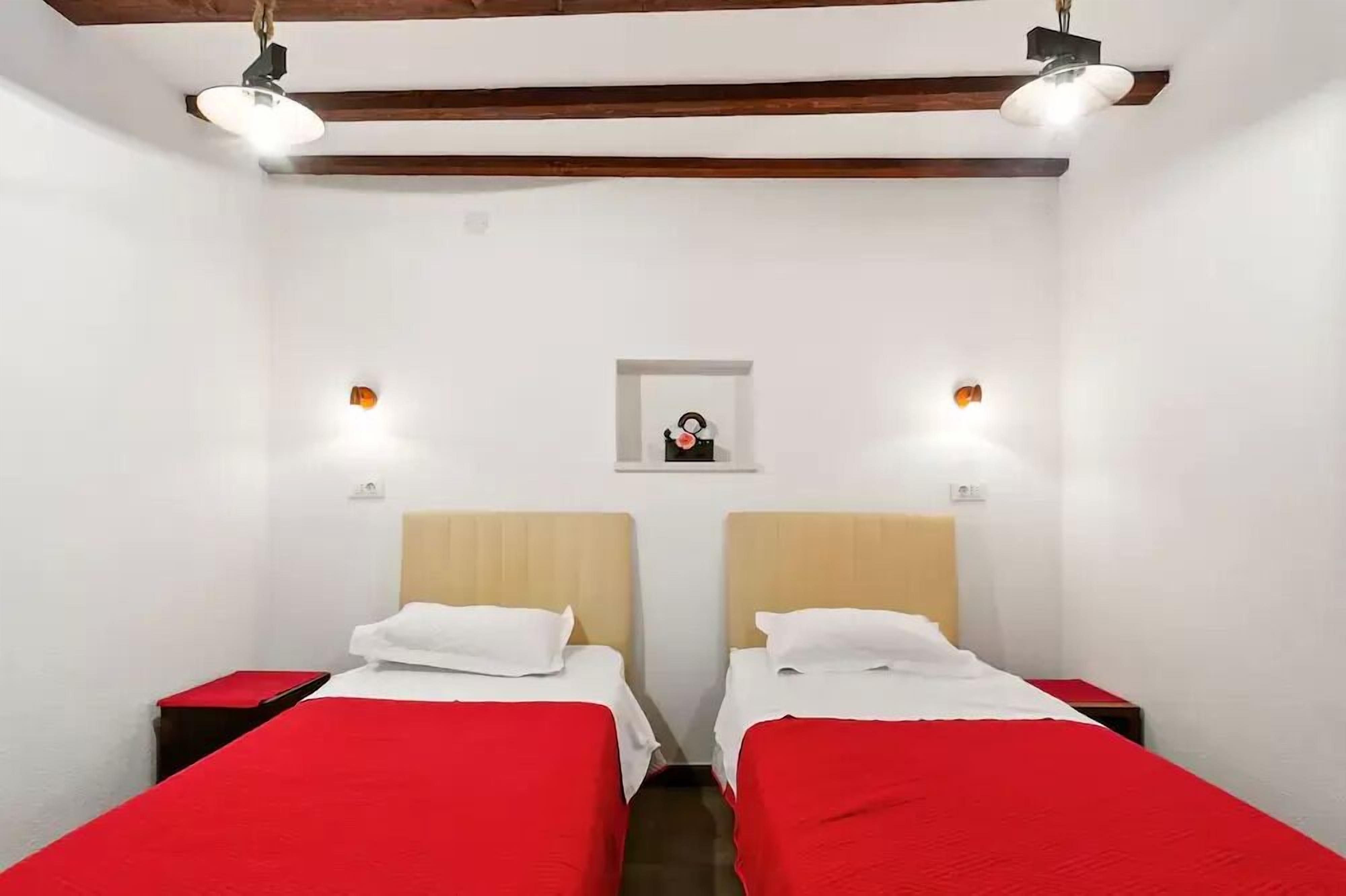 A room with two beds, each with a red bedspread and white pillows, and a small window with a picture on the wall.