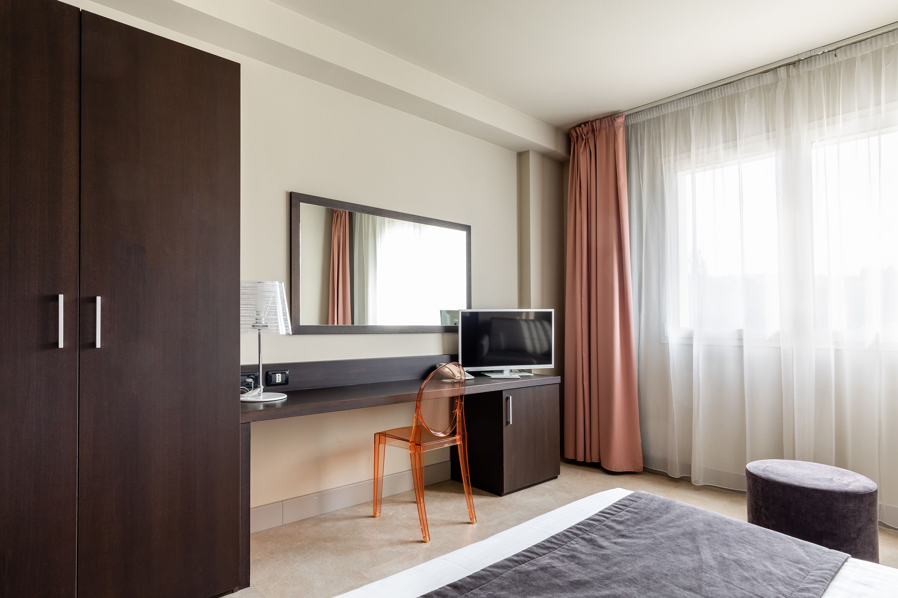 Una habitación de hotel moderna con una cama grande, un escritorio con computadora, una silla, un televisor y un ventanal con cortinas.