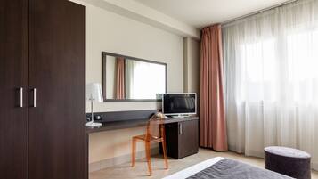 Una habitación de hotel moderna con una cama grande, un escritorio con computadora, una silla, un televisor y un ventanal con cortinas.