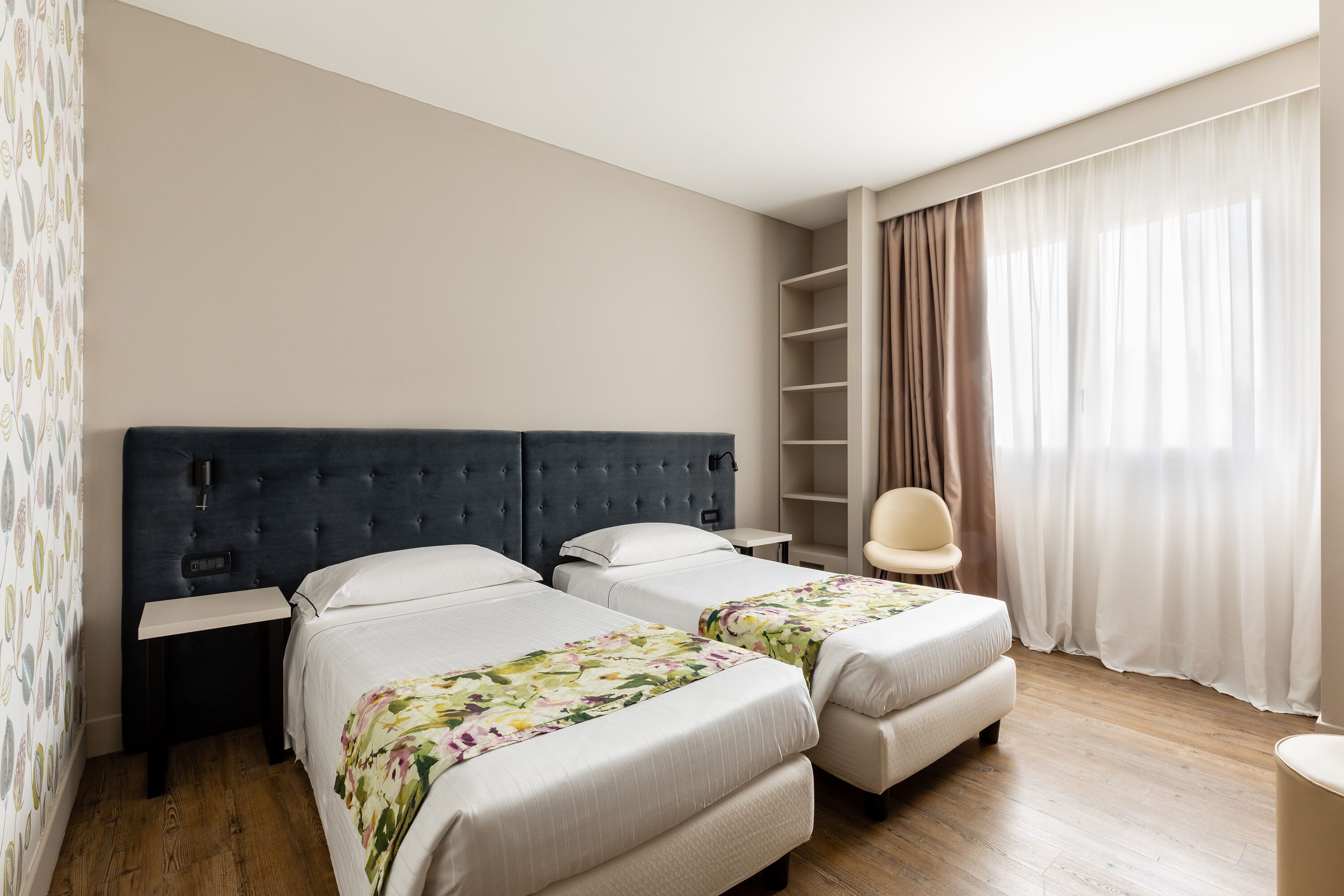 Ein Hotelzimmer mit zwei Betten, einem Nachttisch, einem Sessel, einem Bücherregal und einem großen Fenster mit Vorhängen.