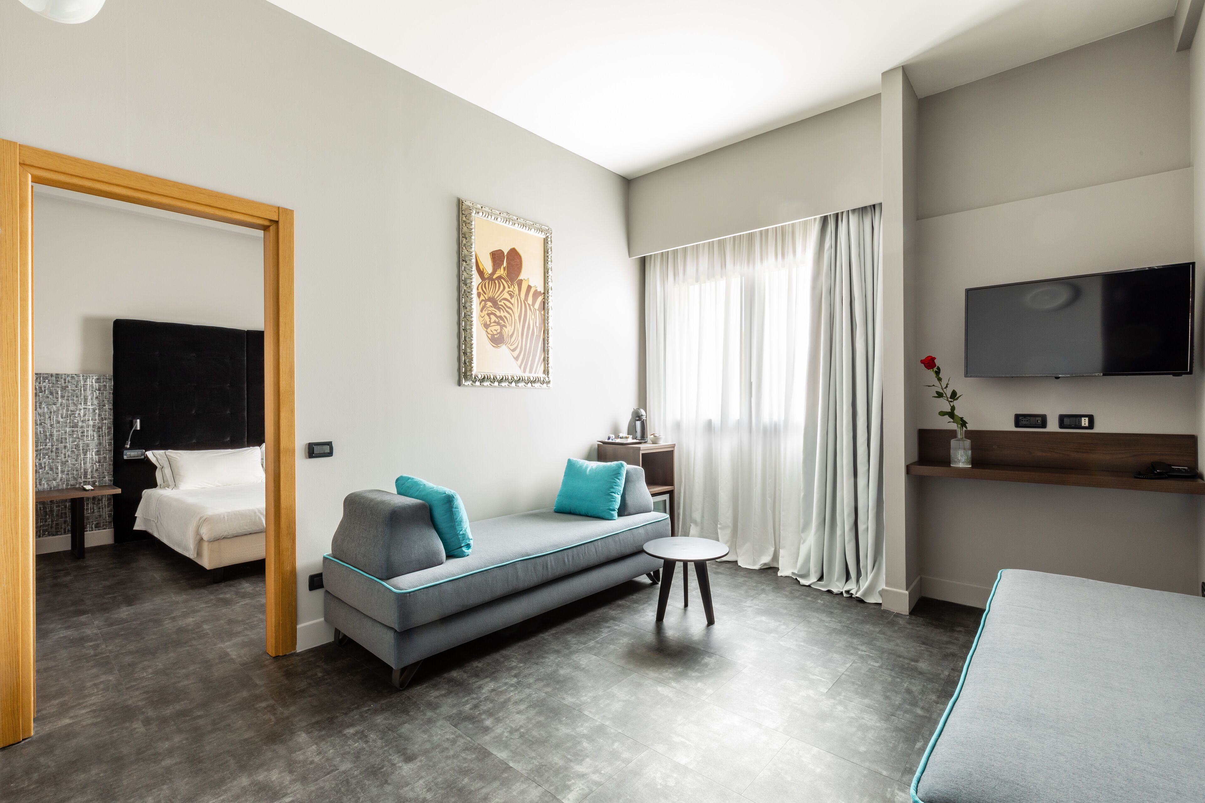 Una habitación de hotel moderna con un sofá gris, una mesita redonda pequeña, una cama, un televisor montado en la pared y una obra de arte enmarcada sobre el sofá.
