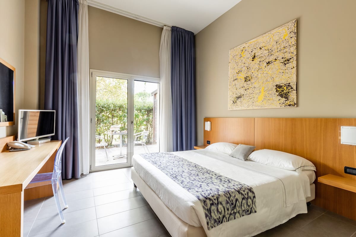 Ein Hotelzimmer mit Bett, Schreibtisch mit Computer, Fernseher und einem großen Fenster mit Vorhängen.