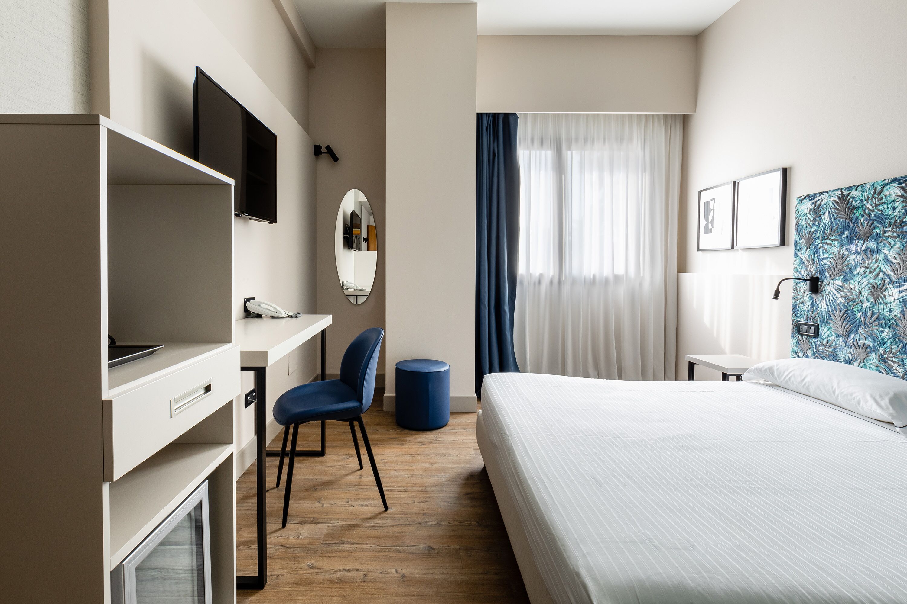 A hotel room with a bed, desk, chair, TV, and a large window with curtains.
