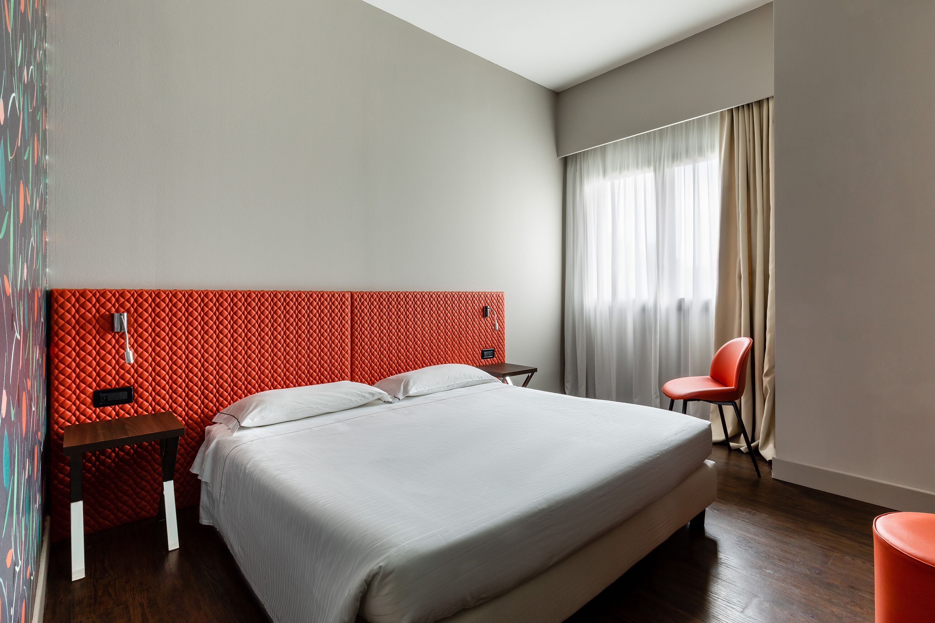 Una habitación de hotel moderna con una cama grande, una pared de acento roja, una mesita pequeña, una silla y un ventanal con cortinas.